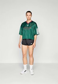 T-shirt vert Adidas raccourci avec col rouge et logo, associé à un short noir. Le modèle porte des baskets blanches et des chaussettes de sport.