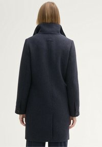 Mujer con cabello lacio castaño claro, vistiendo un abrigo oscuro de color marino con cuello levantado, de pie, de espaldas, contra un fondo claro y liso.