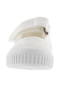 Zapatilla de lona blanca con cinta de velcro, suela de goma texturizada y puntera redondeada. Cuenta con un lazo de metal para facilitar el ajuste del cierre.