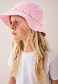 Junges Mädchen mit langen blonden Haaren, das einen pinkfarbenen Fischerhut mit pastellfarbenen Perlen trägt und ein schlichtes weißes T-Shirt anhat, blickt nach links.