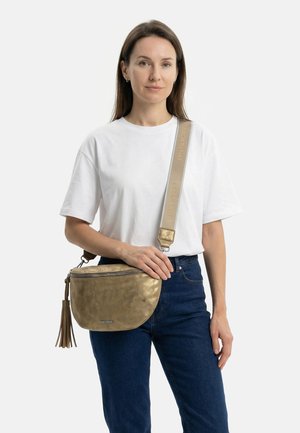 Woman wearing white t-shirt and blue jeans holds gold crossbody bag with wide beige strap and tassel detail.