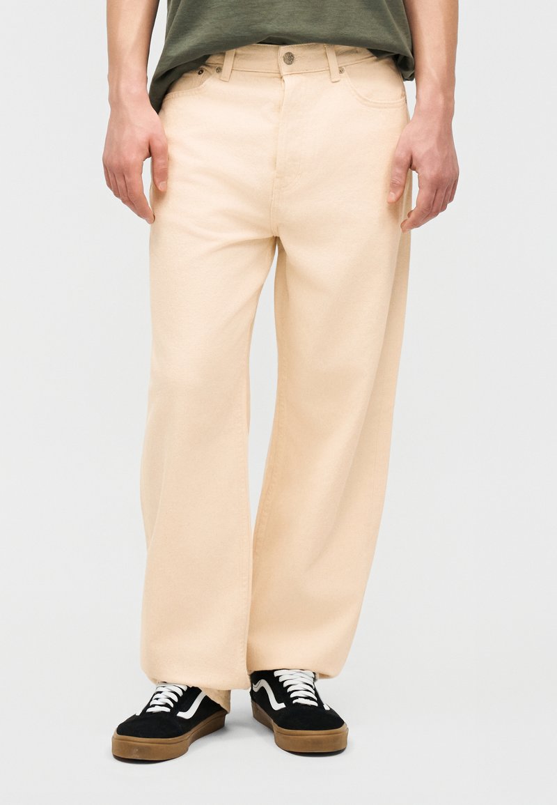 Person iført cremelignende bukser med vide ben, kombineret med sorte og brune sneakers og en grøn skjorte.