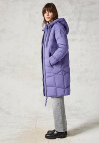 Cappotto imbottito viola con cappuccio, caratterizzato da un design trapuntato, chiusura frontale con zip e tasche laterali. Abbinato a jeans grigi e stivali neri.