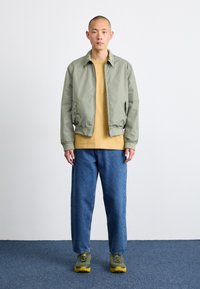 Hellgrüne Bomberjacke, gelbes T-Shirt, lässige blaue Jeans und gr�üne Sneakers mit gelben Akzenten. Einfaches Design und lässige Passform.