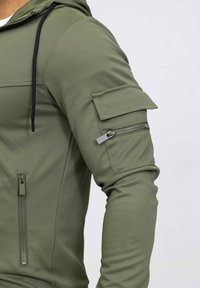 Haut à capuche vert olive en tissu texturé, doté d'une poche zippée sur la manche et d'une capuche à cordon. Design ajusté avec une silhouette élégante.