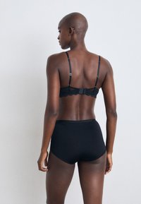 Bralette en dentelle noire avec bretelles ajustables et fermeture à crochets, associé à une culotte noire taille haute, vu de dos.