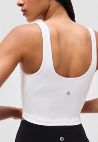 Frau von hinten gesehen, trägt ein weißes kurzes Top und schwarze Leggings, beide mit kleinen reflektierenden Lululemon-Logos.