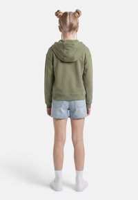 Dziecko o blond włosach związanych w dwa koki, ubrane w oliwkowozieloną bluzę z kapturem, jasne szorty dżinsowe, białe skarpetki i białe trampki, stojące tyłem.