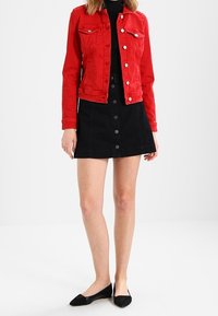 Rote Denimjacke mit Knopfverschluss und zwei Brusttaschen, kombiniert mit einem schwarzen A-Linien-Rock mit vorderen Knöpfen und schwarzen flachen Schuhen.