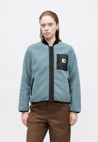 Világoskék polárpulóver fekete zipzárral és zsebbe varrt mellzsebbel. Texturált felszínnel és oldalzsebekkel rendelkezik, kerek nyakkivágással.