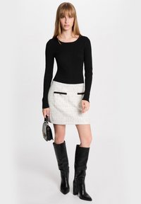 Haut noir à manches longues côtelé associé à une mini-jupe blanche texturée avec des poches noires à passepoil et des bottes noires à talons hauts jusqu'aux genoux.