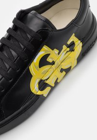 Black leather sneaker with yellow and gray logo detail on the side, black laces, and a smooth texture. Rubber sole visible at the bottom.