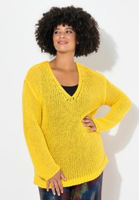 Gelbes V-Ausschnitt gestricktes Sweatshirt im lockeren Schnitt mit langen Ärmeln und strukturiertem Stoff. Kombiniert mit mehrfarbigen gemusterten Hosen.