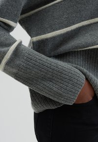 Pull en rayures grises avec texture côtelée ; présente des lignes horizontales crème et un poignet serré. Porté par-dessus des vêtements noirs.