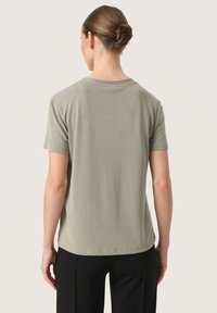 T-shirt a maniche corte, con collo giro, di colore verde oliva tenue, caratterizzato da una texture liscia e morbida e una vestibilità rilassata, abbinato a pantaloni neri.