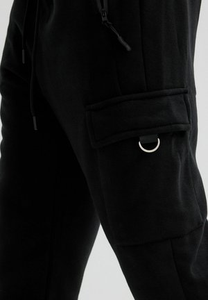 Pantalones negros con un bolsillo con solapa en el muslo que presenta un detalle de anilla en D metálica y un cordón ajustable en la cintura.