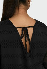 Blusa nera caratterizzata da un retro a V con chiusura a lacci, tessuto texture e motivi a pois su tutta la superficie. Le maniche sono trasparenti.