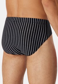 Slip de bain noir avec des rayures blanches verticales, en tissu doux, offrant un ajustement confortable et mettant en valeur un design minimaliste avec une taille mi-haute.
