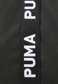 Tissu noir avec un logo "PUMA" blanc bien visible, présentant une surface texturée et des coutures solides. Accent sur le matériau et le détail du logo.