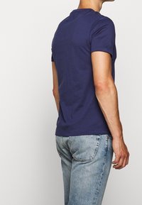 Muže v navy modrém tričku s krátkým rukávem a světle modrých džínách, stojící zády a z pravé strany viditelný na jednobarevném pozadí.