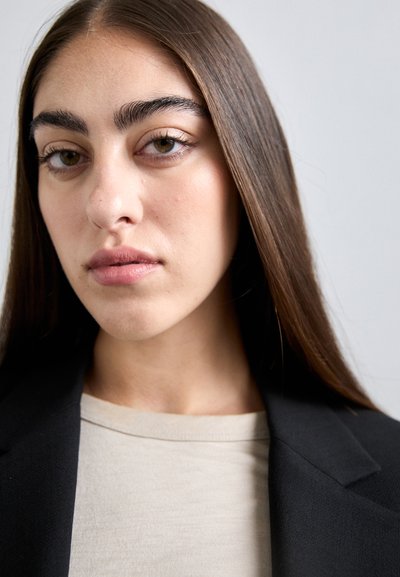 Close-up of a face, featuring long brown hair, straight brows, and neutral lips. The subject wears a beige top and a black blazer.