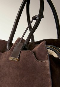 Bruine suède handtas met leren hengsels, voorzien van een gouden hardwareaccent en een ritsdetail. Gecoate oppervlakte met gestikte randen.