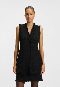 Schwarzes Tweed ärmelloses Kleid mit strukturiertem Finish, V-Ausschnitt und vier Knöpfen auf der Vorderseite. Kurzer Schnitt mit Fransenverzierung.