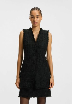 Mustat tweed-viittamaton mekko, jossa on tekstuuripinta, V-kaula-aukko ja neljä nappia edessä. Lyhyt malli, jossa on tupsutuksia.