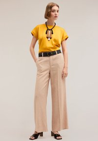 Żółta bluzka z krótkim rękawem, beżowe szerokie spodnie w paski, czarny pleciony pasek z okrągłą klamrą oraz czarne sandały na słupku.