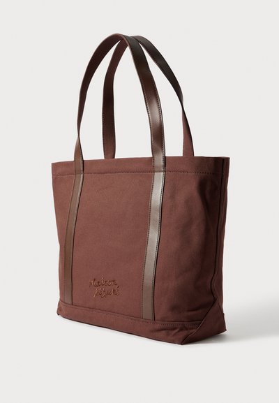 Barna vászontáska sötétbarna bőr fogantyúkkal és szegéllyel, a "Maison Kitsuné" hímzéssel az eleje bal alsó oldalán.