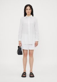 Camicia bianca a maniche lunghe, con bottoni frontali, abbinata a pantaloni corti bianchi. Una borsa nera e sandali neri completano l'outfit.