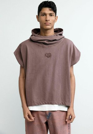 Młody mężczyzna ubrany w luźną brązową bezrękawnikową bluzę z kapturem z emblematem serca, białą koszulkę pod spodem oraz wyblakłe różowe dżinsy, stojący na jednolitym tle.