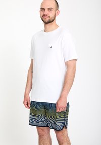 Homme barbu portant un t-shirt blanc uni à manches courtes et un short à motifs bleu-vert, debout devant un fond blanc uni.