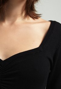 Haut noir en tricot avec un décolleté en V profond, présentant des fronces au centre et des bords côtelés texturés sur les manches.
