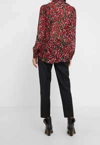Blusa negra transparente con estampado floral en rojo y blanco, de mangas largas, con puños fruncidos y un corte holgado, combinada con pantalones negros entallados y botas hasta el tobillo.