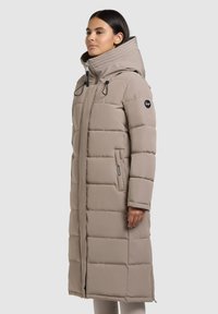 Beiger gepolsterter Mantel mit hohem Kragen und Kapuze, versehen mit schwarzen Akzenten, Reißverschluss vorne und seitlichen Taschen, entworfen für Wärme und Komfort.