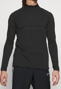 Schwarzes, langärmliges Sportoberteil mit einem Viertelreißverschluss, das eine glatte Stofftextur und ein tonales Paneeldesign über der Brust aufweist.