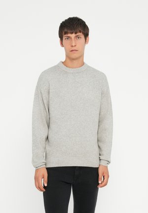 Light gray knit sweater with a round neck, dropped shoulders, and ribbed cuffs and hem. Paired with black jeans, model stands against a white background.