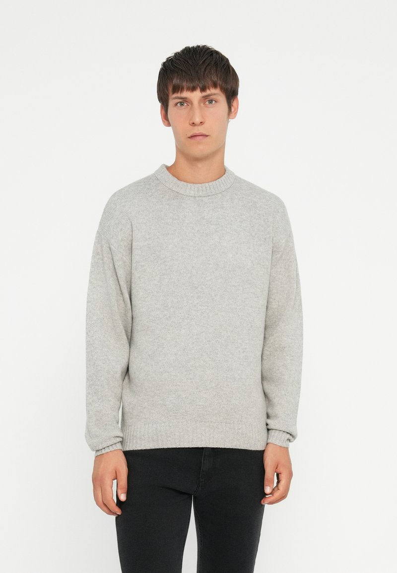 Light gray knit sweater with a round neck, dropped shoulders, and ribbed cuffs and hem. Paired with black jeans, model stands against a white background.
