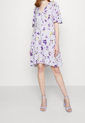 Kvinde iført en knælængde, lys lilla blomsterkjole med korte ærmer og matchende lavendel høje hæle sandaler, stående på en ensfarvet baggrund.