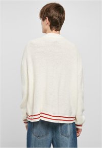 Megzta svetaine off-white spalvos, laisvas kirpimas ir raudonais dryžiais dekoruota juosta apatinėje dalyje. Turi ilgomis rankovėmis ir tekstūruotą, atvirą audinį.