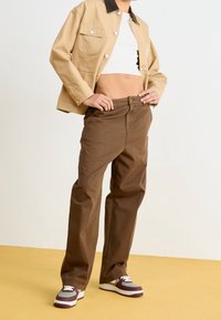 Veste en coton beige avec un col contrastant, haut court blanc, pantalon droit marron, et baskets multicolores ; texture lisse.