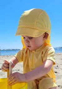 Gul, kortærmet solbeskyttelsesoutfit med lynlås. En matchende kasket dækker hovedet. Barnet leger med en gul spand i sandet.