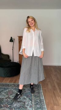 Γυναίκα με ξανθά μαλλιά και χαμόγελο φοράει λευκό πουκάμισο, ασπρόμαυρη καρό φούστα, μαύρες κάλτσες και μαύρα αθλητικά παπούτσια Adidas, στέκεται σε χαλί με σχέδια.