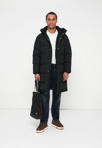 Sort pufferjakke med hætte og forreste knapper, parret med en hvid skjorte, mørke jeans, brune sneakers og en sort tote bag.