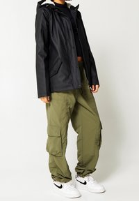 Schwarze isolierte Jacke mit Kapuze, ausgestattet mit einem Frontreißverschluss und Druckknopfverschluss. Kombiniert mit lockeren olivgrünen Cargo-Hosen und weißen Sneakers.