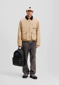 Beige jakke med glidelås og frontlommer, svart krage, sammen med grå jeans. Svart ryggsekk med blå aksent. Beige caps.