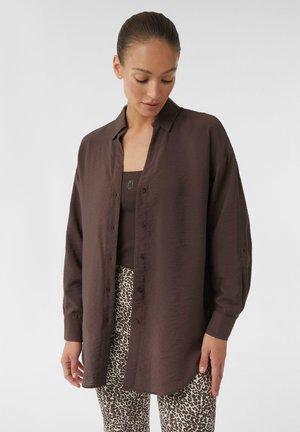 Brown, button-up shirt with a soft, textured finish, featuring a classic collar and long sleeves, worn over a dark tank top and patterned pants.