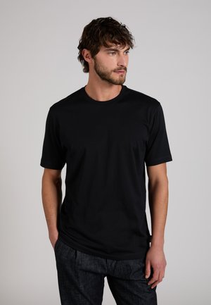 Mann mit Bart und lockigem Haar, trägt ein schwarzes kurzärmeliges T-Shirt und dunkle Hose, schaut nach rechts, die Hände in den Taschen.