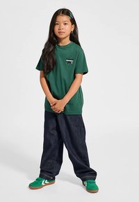 Grön bomullst-shirt med en liten logotyp, löst sittande mörka denimbyxor och gröna sneakers med vita detaljer. Stående pose med händerna sammanflätade.